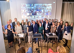 Gruppe von Menschen steht vor einer Wand mit einer Projektion von vielen kleinen Videokonferenzfenstern