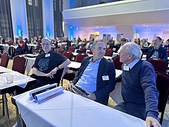 Blick in einen Zuschauerraum, Menschen sitzen hinter- und nebeneinander an Tischen