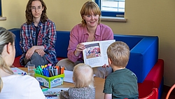 Frau zeigt Kindern ein Buch mit bunten Illustrationen in einem Raum mit bunten Möbeln und einem Tisch mit Stiften
