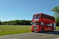 Ein Londonbus fährt auf der Straße, im Hintergrund eine grüne Wiese