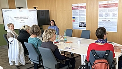 Mehrere Frauen sitzen an einem Konferenztisch in einem Schulungsraum mit Präsentationsfolie und Informationsplakaten an der Wand