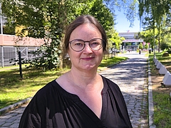 Frau mit Brille und schwarzem Oberteil steht auf einem gepflasterten Weg im Freien mit Bäumen und Gebäuden im Hintergrund