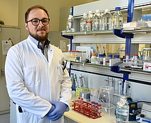 Person mit weißem Laborkittel und blauen Handschuhen steht vor Laborarbeitsplatz mit Reagenzgläsern, Flaschen und Laborgeräten