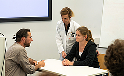 Eine Studentin und ein gestellter Patient sitzen sich am Tisch gegenüber eine Ärzte beugt sich über die Studentin und gibt Hilfestellung