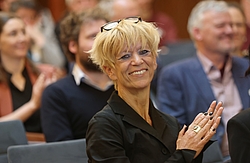 Frau mit kurzen blonden Haaren schaut in die Kamera, im Hintergrund weitere Menschen