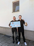 Zwei Männer stehen vor einer weißen Hauswand, einer hält einen großen Scheck mit der Aufschrift 550,- €, der andere eine kleine Plüschfigur