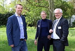 Zwei Männer und eine Frau in dunkler Kleidung stehen im Grünen.