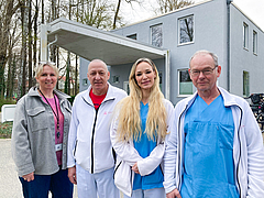 Vier Personen in medizinischer Arbeitskleidung stehen vor einem modernen Gebäude mit großen Fenstern und einem Vordach