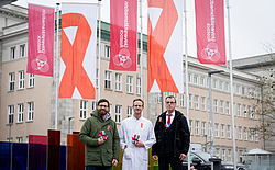 3 Personen stehen nebeneinander vor gehissten Fahnen.