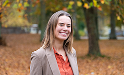Frau mit schulterlangem Haar trägt beige Blazer und orangefarbene Bluse vor herbstlichem Waldhintergrund