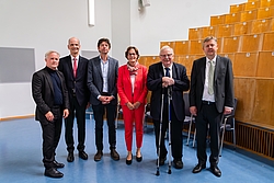 Sechs Personen in formeller Kleidung stehen in einem Hörsaal mit Holzsitzreihen und blauer Bodenfläche nebeneinander