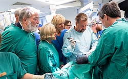 Viele Personen stehen nah beieinander um einen OP-Tisch und schauen gespannt zu.