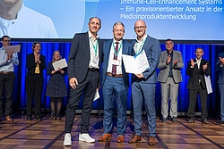 3 Personen stehen nebeneinander auf einer Bühne, lächeln und halten eine Auszeichnung in der Hand. Im Hintergrund klatschen viele andere Personen.