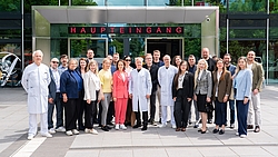 Gruppe von Menschen in Business- und Arztkleidung steht vor einem Gebäude mit der Aufschrift „HAUPTEINGANG"