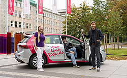 Zwei junge Menschen stehen neben einem farbig beklebten Auto, ein junger Mann sitzt vor geöffnerter Tür auf dem Beifahrersitz