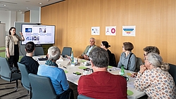 Gruppe von Menschen sitzt an einem Konferenztisch und hört einer Frau zu, die vor einem Bildschirm mit Präsentationsfolien steht.