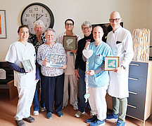 Gruppe von sieben Personen steht in einem Raum und hält gerahmte Klöppelarbeiten und ein langes, herzförmiges Dekorationsstück.