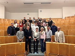 Gruppe von Menschen in einem Hörsaal mit hölzernen Sitzreihen, die in mehreren Reihen auf Treppenstufen stehen und in die Kamera lächeln