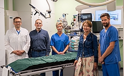 Fünf Personen in unterschiedlicher Krankenhauskleidung und in zivil stehen um eine Trage herum in einem Untersuchungsraum