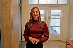 Eine Frau in einer roten Bluse und steht in einem Gang vor einer Tür und lächelt.