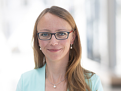 Frau mit langen braunen Haaren, Brille und hellblauem Blazer lächelt in die Kamera vor hellem, unscharfem Hintergrund