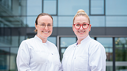 Zwei Frauen in weißen Kitteln stehen vor einem modernen Glasgebäude